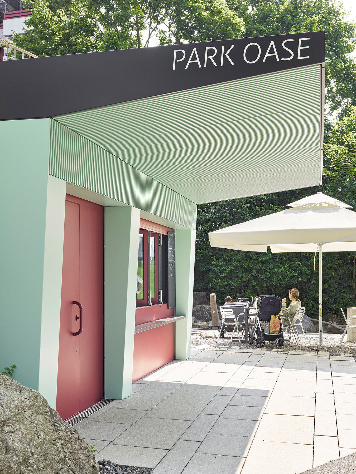 Blick auf die Buvette „Park Oase“ im Kyburgpark Sursee vom Sitzbereich mit Sonnenschirm, roten Fensterrahmen, grünem Stahlvordach und Terrasse.
